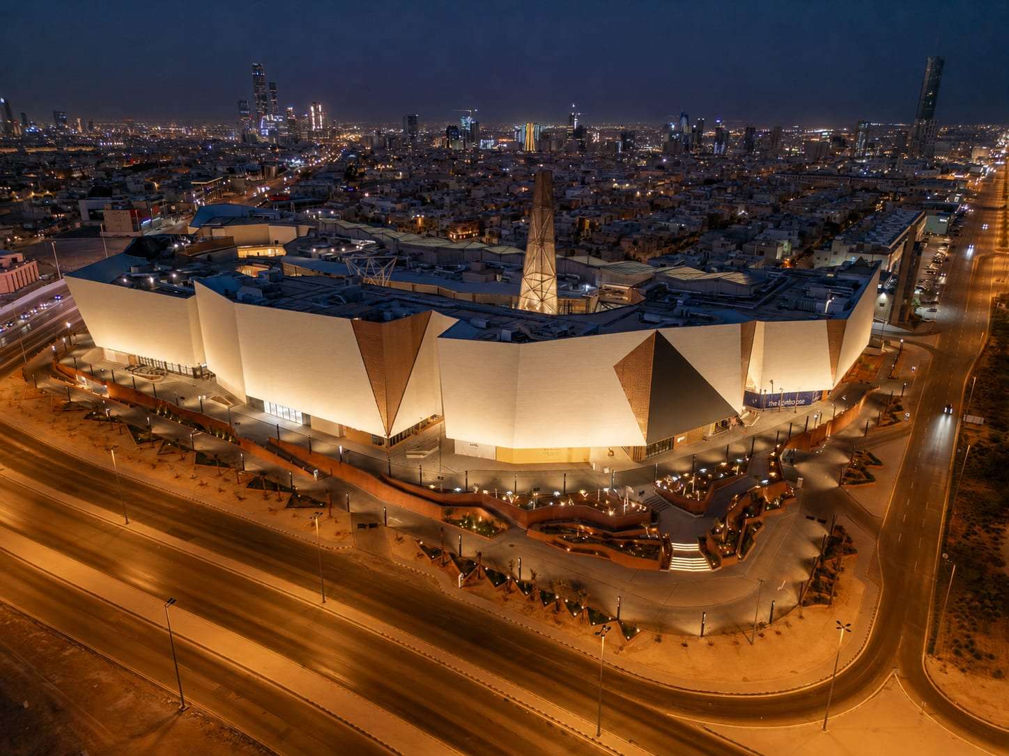 A retail and leisure block seen from above at dusk, each facade plane lit to its own value against the city behind it